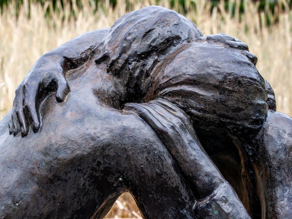 Darstellung zweier kniender Menschen, die sich innig in den Armen liegen. Unter bzw. zwischen ihnen liegt eine Bibel und ein Stück Stacheldraht. Die Bronzefiguren sind auf einem schmalen quaderförmigen Sockel aus Beton platziert, der mit Steinen besetzt ist.