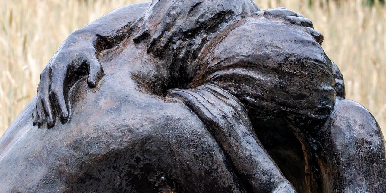 Darstellung zweier kniender Menschen, die sich innig in den Armen liegen. Unter bzw. zwischen ihnen liegt eine Bibel und ein Stück Stacheldraht. Die Bronzefiguren sind auf einem schmalen quaderförmigen Sockel aus Beton platziert, der mit Steinen besetzt ist.