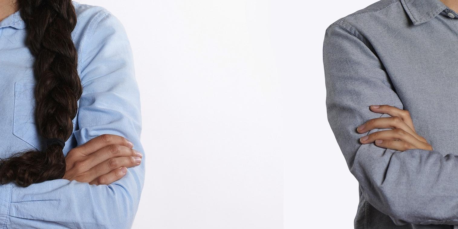 Zwei Menschen stehen mit verschränkten Armen mit größerem Abstand nebeneinander. Man sieht, dass es eine Frau (links) und ein Mann (rechts) ist, aber kein Gesicht.