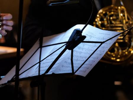 Notenständer, auf dem Notenblätter liegen, von hinten fotografiert, dahinter im Ausschnitt zwei Musiker mit ihren  Blasinstrumenten.