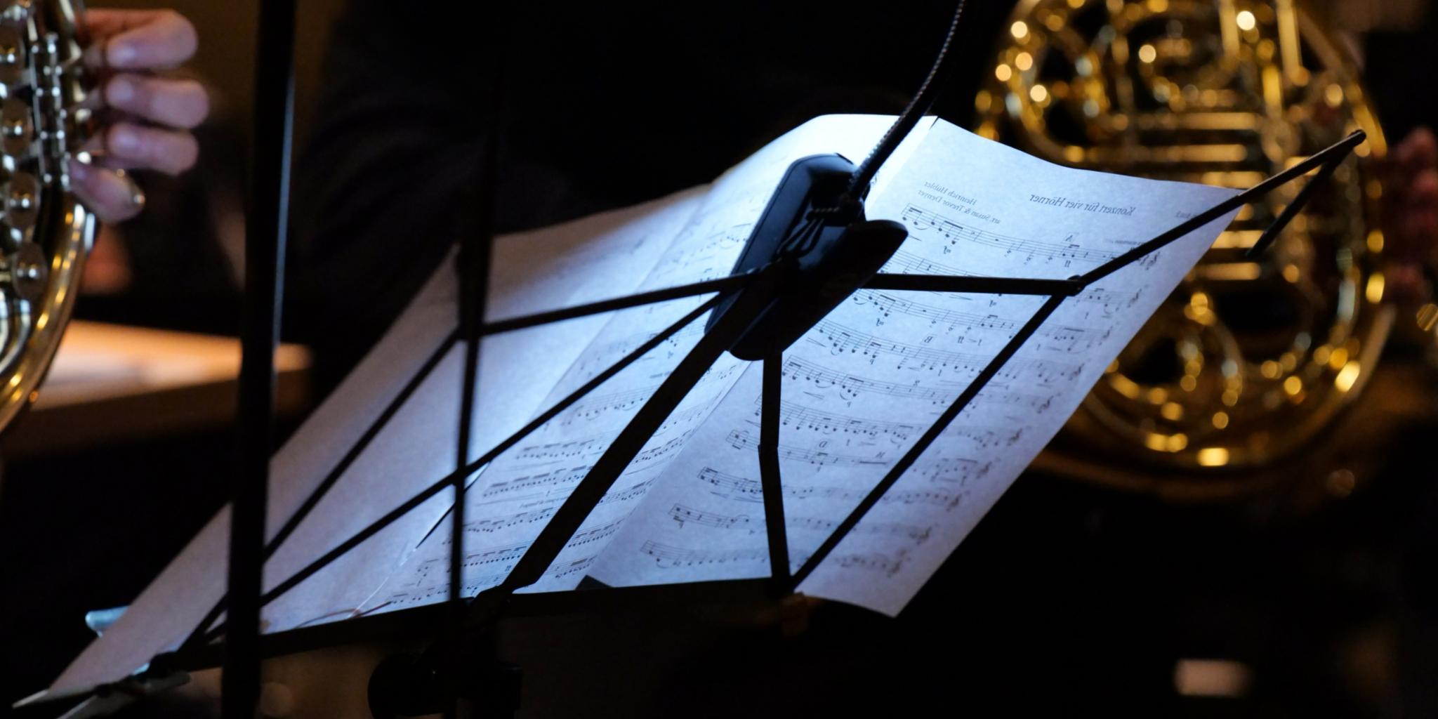 Notenständer, auf dem Notenblätter liegen, von hinten fotografiert, dahinter im Ausschnitt zwei Musiker mit ihren Blasinstrumenten.