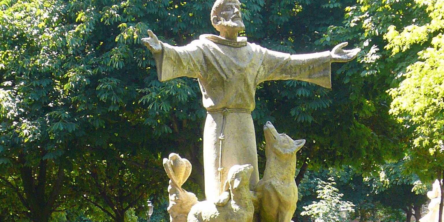 Skulptur des heiligen Franziskus in einem Park vor Bäumen, neben ihm stehen ein Wolf und ein Lamm. Der Heilige hat die Arme weit ausgebreitet.