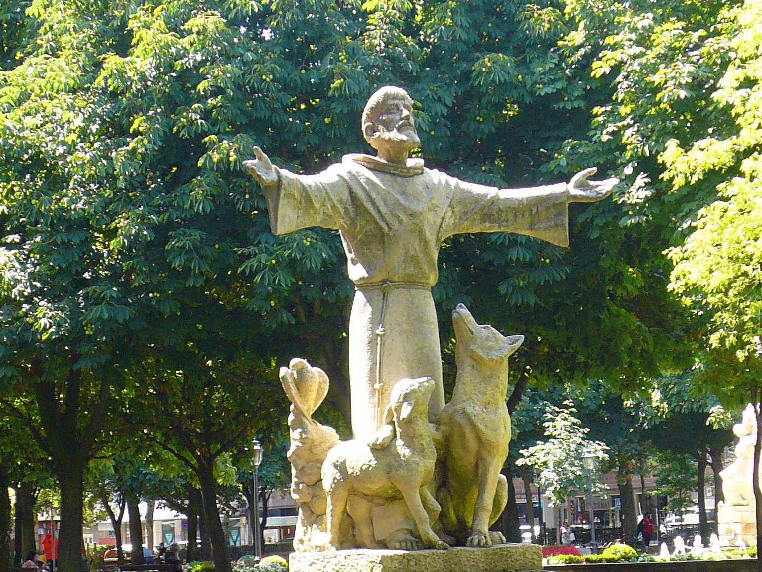 Skulptur des heiligen Franziskus in einem Park vor Bäumen, neben ihm stehen ein Wolf und ein Lamm. Der Heilige hat die Arme weit ausgebreitet.