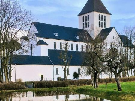 Ein Klostergebäude mit Kirchenschiff und Kirchturm inmitten eines Gartens mit einem kleinen Weiher davor.
