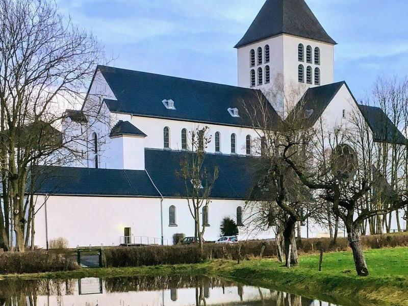 Ein Klostergebäude mit Kirchenschiff und Kirchturm inmitten eines Gartens mit einem kleinen Weiher davor.