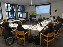 Junge wissenschaftlich Tätige sitzen an Tischen und hören einem Vortrag zu.