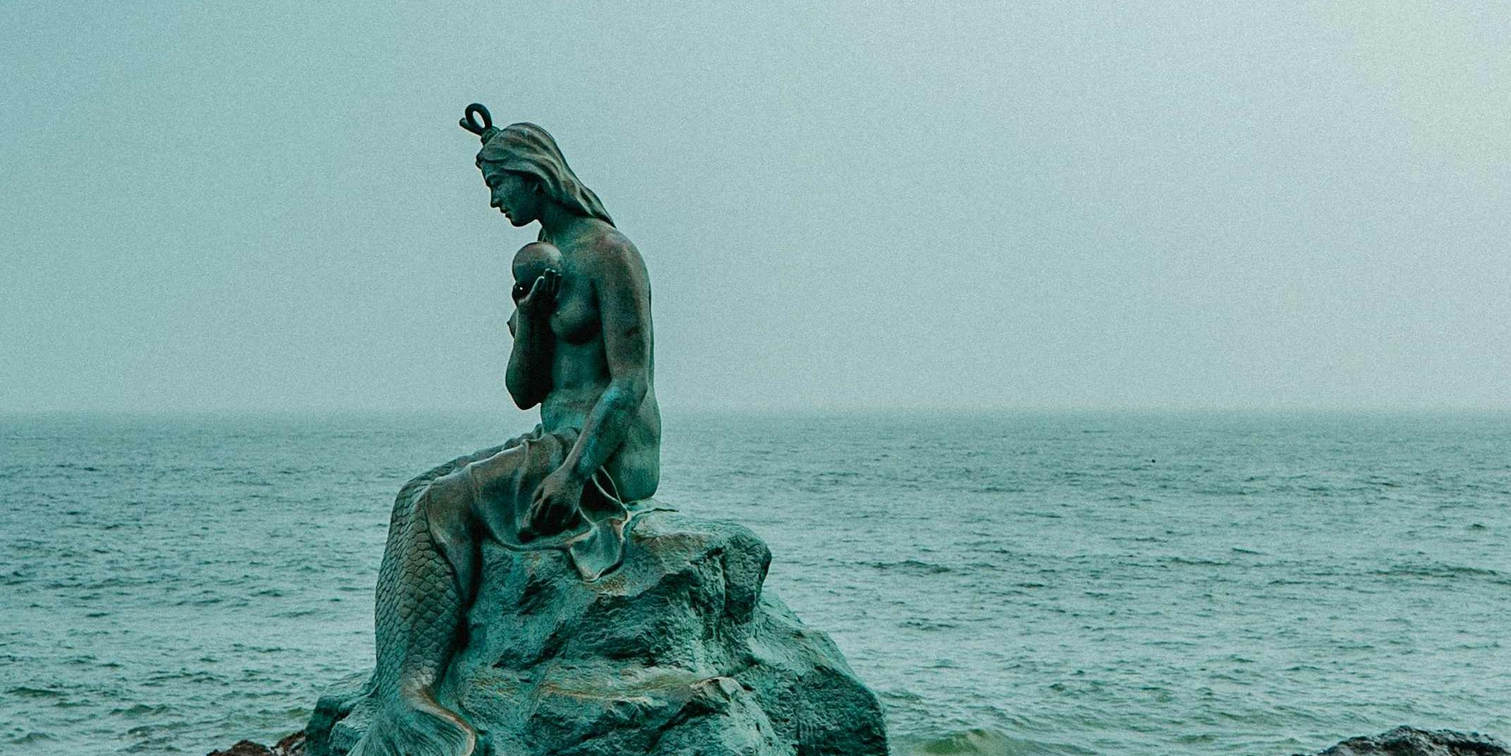 Skulptur einer Meerjungfrau, die traurig auf einem Stein sitzt und eine große Perle