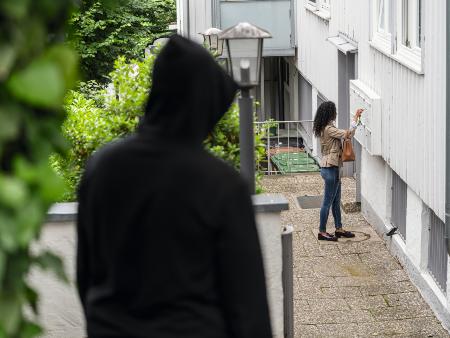 Frau steht an ihrer Wohnungstür und öffnet dort den Briefkasten, während ein Mann sie beobachtet