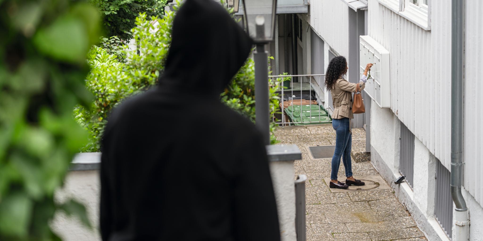 Frau steht an ihrer Wohnungstür und öffnet dort den Briefkasten, während ein Mann sie beobachtet