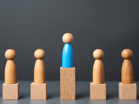 Fünf Holzspielfiguren auf kleinen Holzpodesten, die mittlere Figur steht auf einem höheren Podest, sie überragt die anderen.