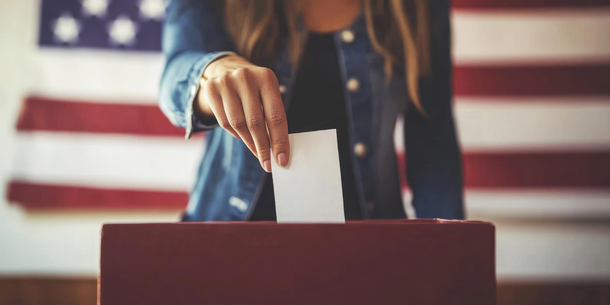 Eine Frau steckt einen Wahlzettel in eine Wahlurne, das Gesicht ist nicht erkennbar. Hinter ihr die Flagge der USA.